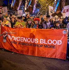 photo of people, some in traditional clothing, at a march carrying a large banner reading: Indigenous Blood, not a single drop more
