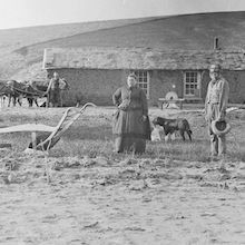 historical black and white photo of two farmers with a plow in front of a farmhouse