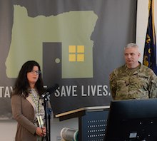 photo of two people at a press conference in front of a banner with the outline of Oregon