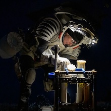 photo taken in the dark of person in mock spacesuit bending over some scientific equipment